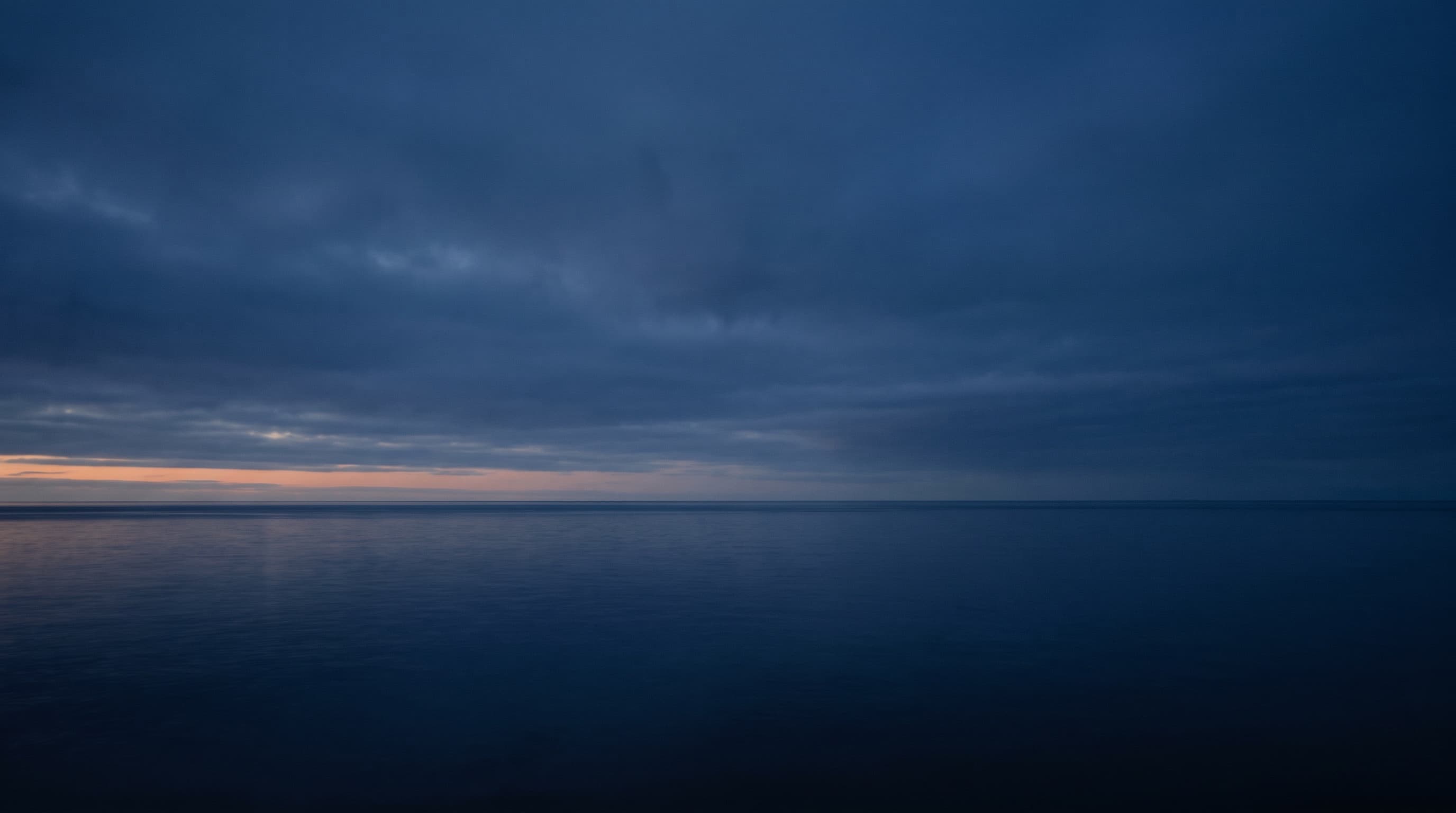 A vast calm sea under a moody deep-indigo dusk sky, a low band of warm light far on the horizon.